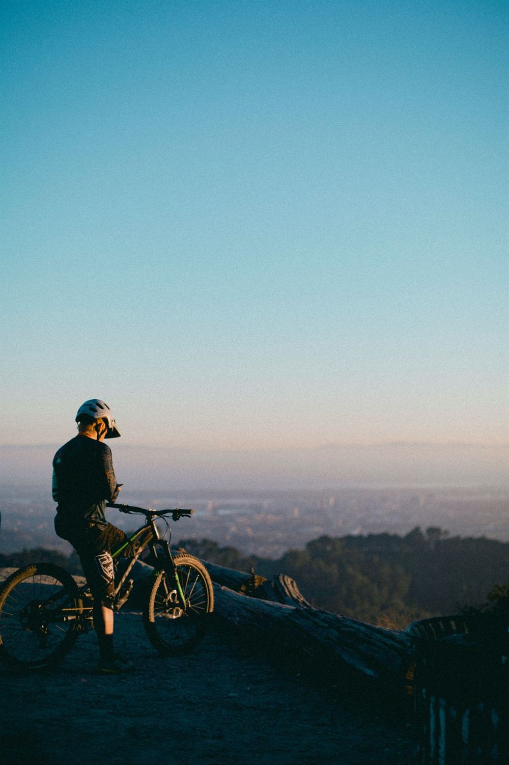A mountain biker at a viewpoint above the city at sunset, looking at the phone